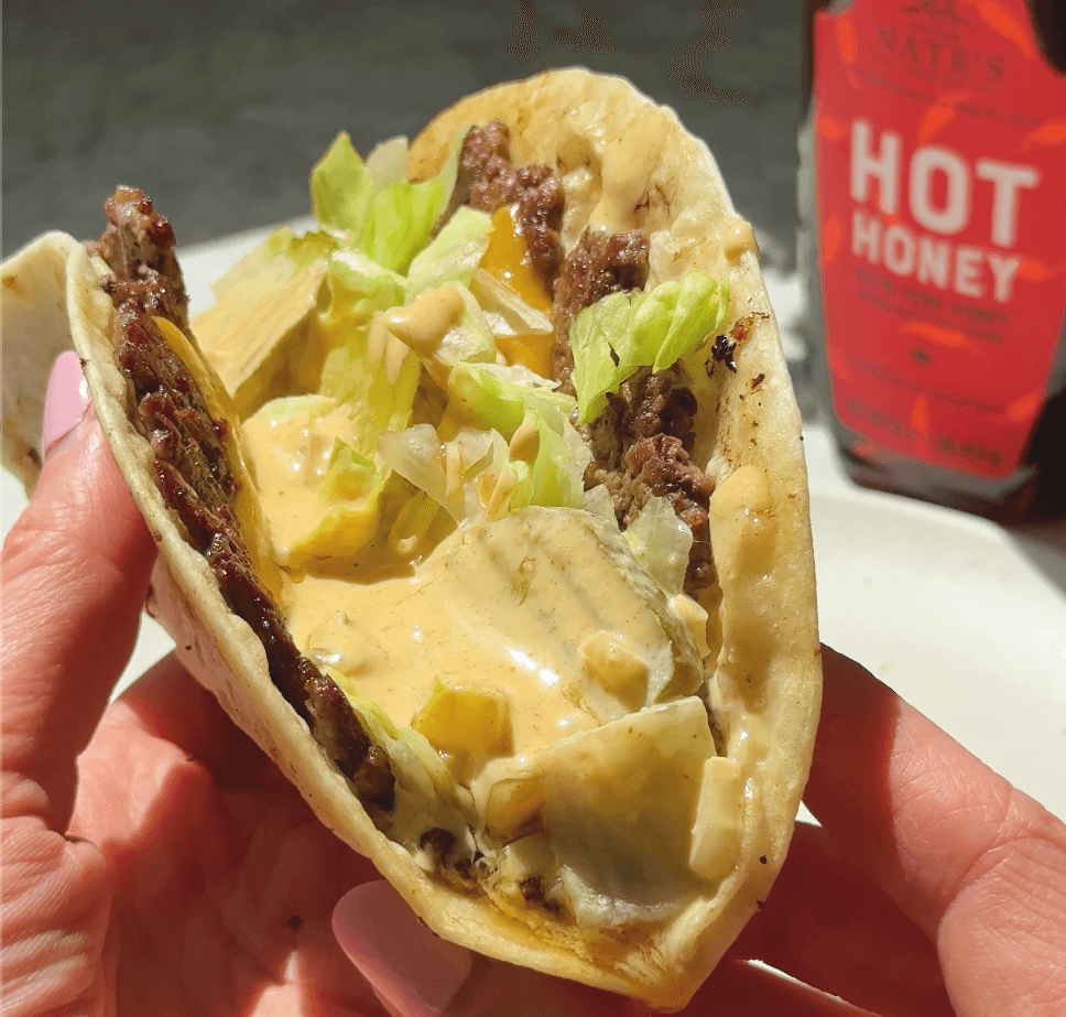 Close-up of a hand holding a burger taco filled with ground beef, chopped lettuce, pickles, and a creamy orange sauce, with a bottle of Nate’s Hot Honey in the background.